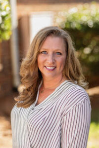 Angela Fisher, a woman with long hair, wearing a striped shirt, smiles at the camera.